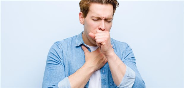 Junger Mann mit rotem Kopf, der sich mit Halsschmerzen und Grippesymptomen krank fühlt, hustet mit zugehaltenem Mund.