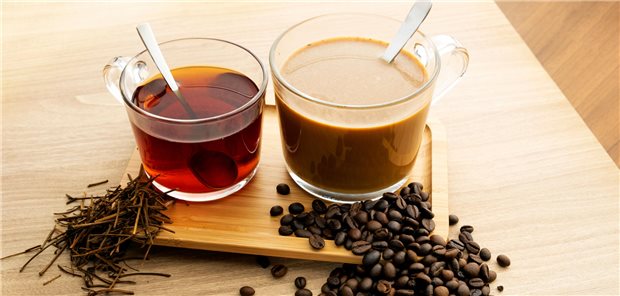 Eine Tasse mit heißem schwarzem Tee und eine Tasse mit heißem Kaffee mit Milch und neben den Tassen die Zutaten für die Heißgetränke Teeblätter und Kaffeebohnen.