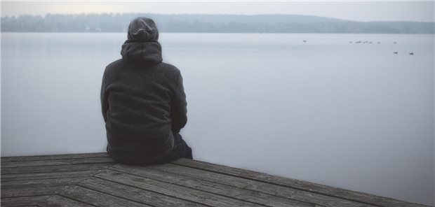 Eine Person sitzt mit dem Rücken zur Kamera an einem See im Nebel.