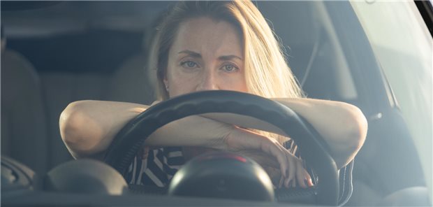 Eine Frau stützt sich mit beiden Armen auf das Lenkrad ihres Autos und legt den Kopf auf den Armen ab.