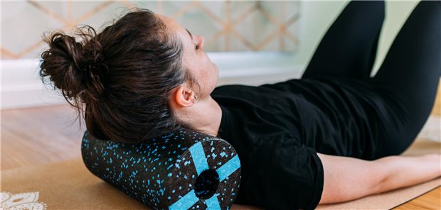 Nackenschmerzen nach Bandscheibenvorfall: Muskeltraining kann helfen Frau in schwarzer Sportkleidung liegt mit einer Faszienrolle unter dem Nacken auf einer Yogamatte und macht Übungen zur Stärkung der Nackenmuskulatur.