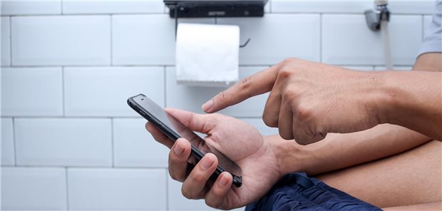Langes Sitzen auf dem Klo erhöht wohl das Risiko für Hämorrhoiden Nahaufnahme einer hand, die am Smartphone tippt. Im Hintergrund sieht man eine Klopapierrolle hängen.