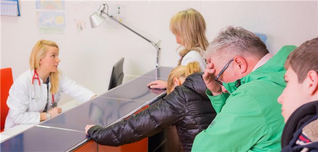 Eine Ärztin sitzt hinter dem Anmeldepult. Es warten viele Patientinnnen und Patienten in einer Schlange. 