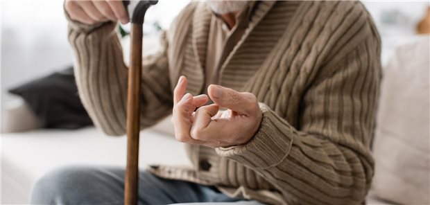 Parkinsonkrankheit: Wie Lebensstil-Interventionen krankheitsmodifizierend wirken Ein älterer Mann sitzt auf einer Couch mit seinem Gehstock. Seine linke Hand hält er gekrümmt.