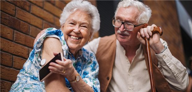 Ein altes Paar sitzt vor einer Mauer. Die Frau hat einen Zuckersensor am Arm und hält ihr Smartphone an den Sensor.