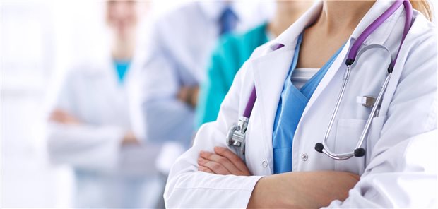 Eine Frau in einem weißen Arztkittel mit verschränkten Armen im Vordergrund. Im Hintergrund sind Kolleginnen und Kollegen zu sehen.