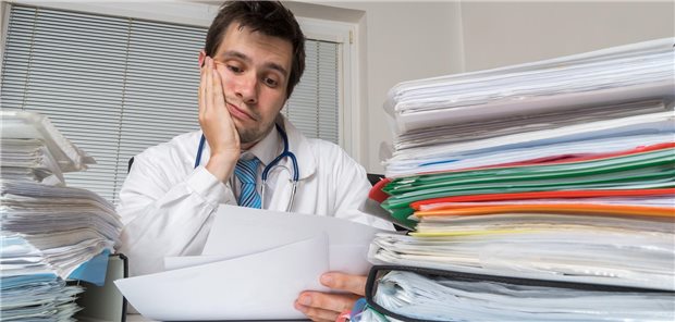 Ein Arzt schaut genervt auf Blätter, während dicke Aktenordnet um ihm herum liegen.